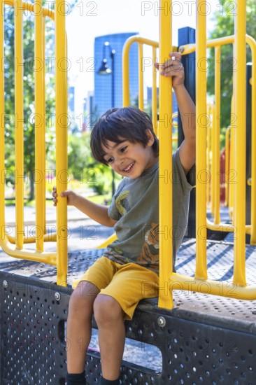 Young boy with a joyful expression sits on a playground structure, holding onto yellow bars and smiling, enjoying a sunny day in a city park in calgary, canada