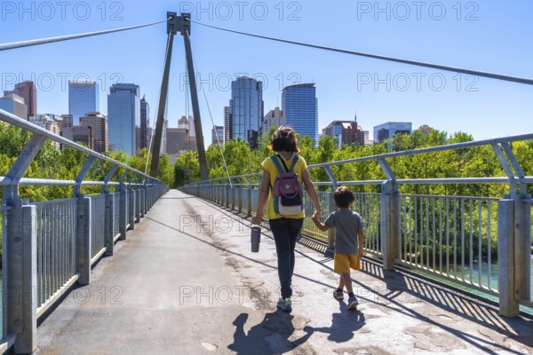 Mother and child are enjoying a leisurely stroll across a pedestrian bridge, set against the backdrop of the calgary skyline on a sunny day