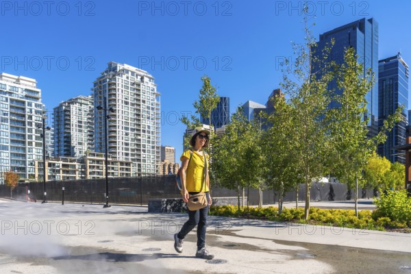 Tourist wearing a yellow t shirt and a hat is enjoying a walk in calgary's east village neighborhood on a sunny day, with modern residential buildings and skyscrapers in the background