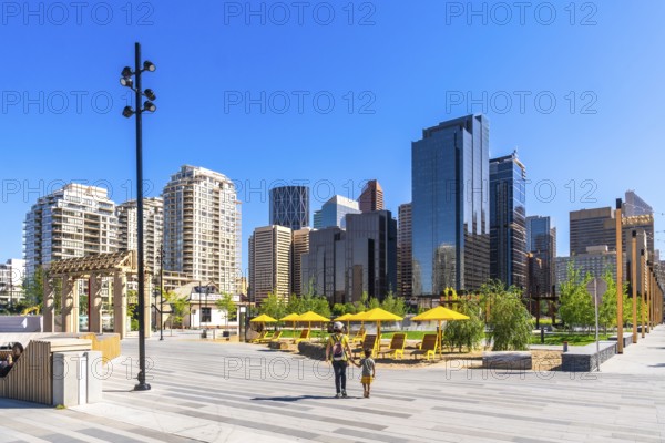 Modern skyscrapers rise above a newly developed urban park in calgary's east village, where a parent and child are enjoying a stroll on a sunny summer day