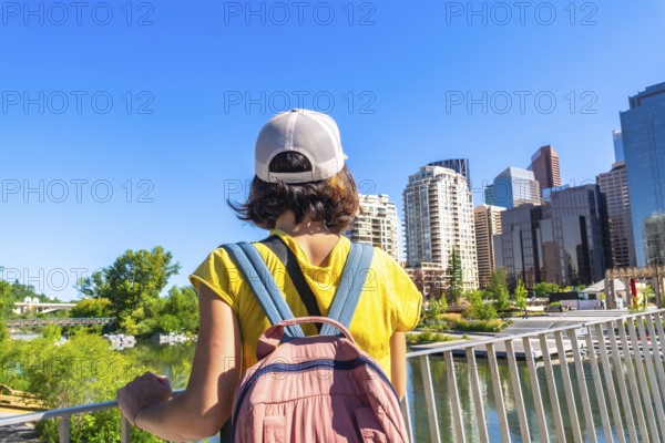 Young woman with backpack standing on peace bridge, admiring modern architecture and cityscape of calgary, alberta, canada, enjoying vibrant urban scenery on bright summer day