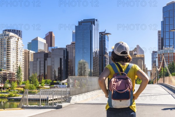 Young woman tourist with backpack admiring the modern skyline of calgary, alberta, canada, on a sunny summer day, standing on a bridge, enjoying urban view