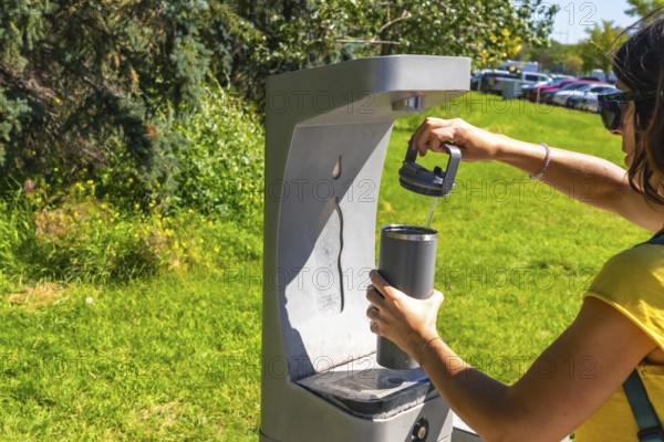 On a sunny day, a tourist is filling her reusable water bottle at a public drinking water fountain in a park in calgary, promoting hydration and sustainability