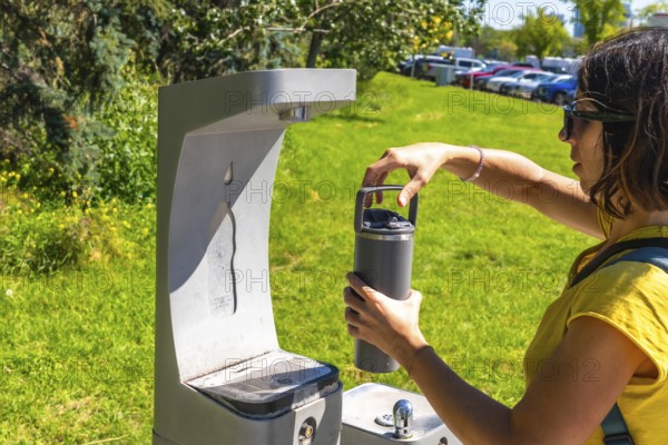 Tourist filling her reusable, eco friendly water bottle at a public drinking water fountain in a park in calgary, promoting sustainability and hydration on a sunny day