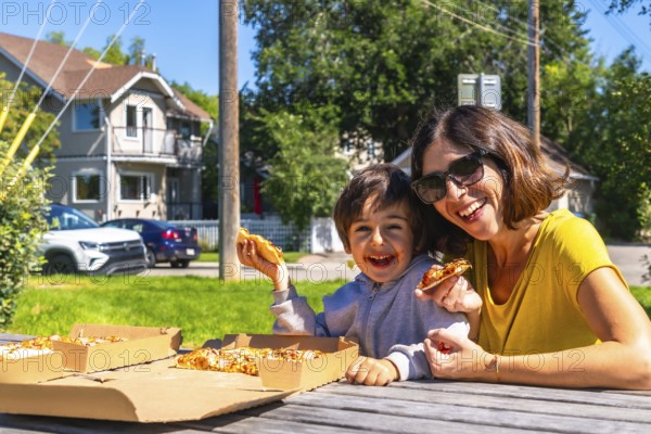 Happy mother and son laughing and eating pizza outdoors at a picnic table in the suburbs of calgary, canada, enjoying quality time together on a sunny day