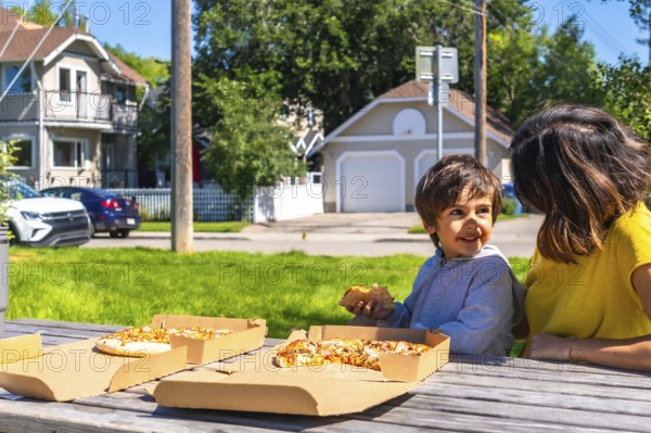 Happy child enjoys a slice of pizza with his mother at a picnic table in a residential neighborhood of calgary, alberta, canada, on a beautiful summer day