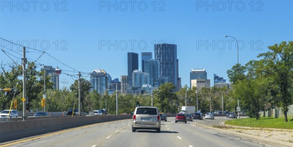 Vehicles are driving on a highway with the calgary skyline, featuring modern skyscrapers and lush greenery, in the background on a clear sunny day