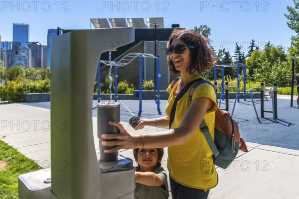Tourist is filling reusable water bottle at a modern public drinking water fountain with calgary skyline in background, promoting sustainability and healthy lifestyle