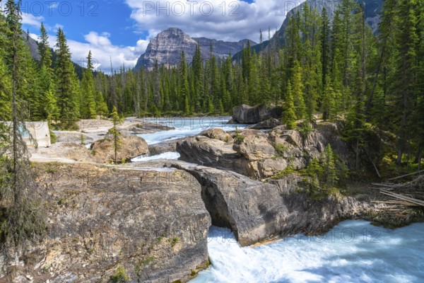 Turquoise bow river flowing beneath a natural bridge rock formation, framed by cascade mountain, pine trees, and a cloudy blue sky in banff national park, alberta