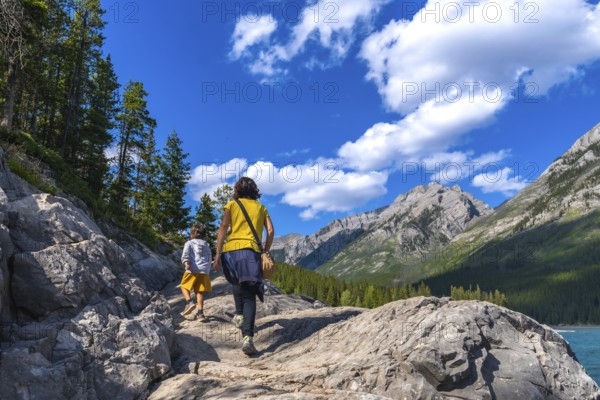 Mother and child hiking on a rocky trail, enjoying the scenic view of lake minnewanka and the surrounding mountains in banff national park, alberta, under a bright blue sky