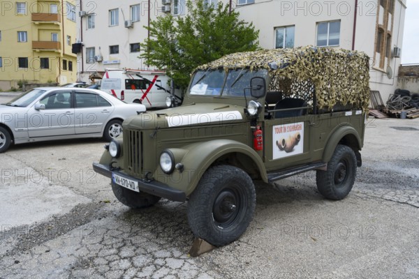 A green vintage car with a camouflage net is parked in a parking lot in front of a building, Soviet off-road vehicle with four-wheel drive, GAZ-69, Georgia