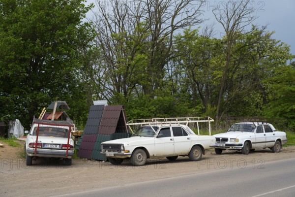 Three older white cars are parked on a rural road next to trees under a cloudy sky, cars with roof racks fixed to the gutter, bumper and a stem, Georgia