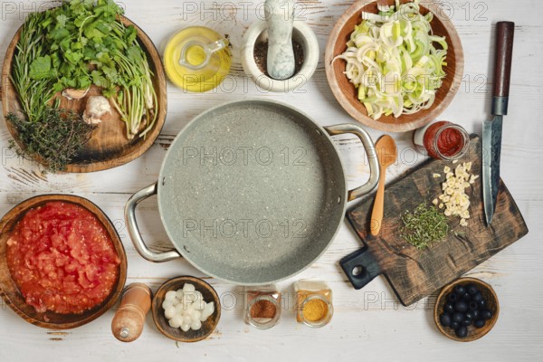 Various food items and cooking tools are arranged on a wooden table. Herbs, vegetables, and spices are present. There is an empty pot in the center ready for cooking