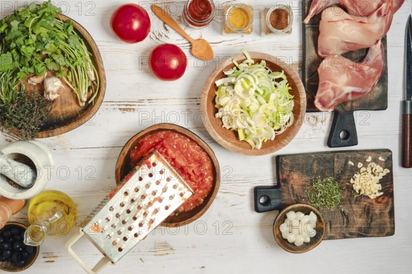 Fresh ingredients are arranged on a wooden table for cooking rabbit stew. There are tomatoes, herbs, garlic, and spices visible. A grater sits nearby, ready for use, and prep looks organized and tidy