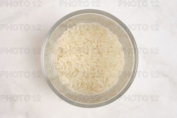 A clear bowl contains uncooked white rice. The rice is fluffy and dry, waiting to be cooked. The bowl is positioned on a light-colored surface, showing a simple kitchen setup