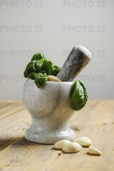 Fresh basil leaves and garlic cloves are placed in a mortar. A pestle is ready to crush the ingredients. The wooden surface adds a natural touch to the scene
