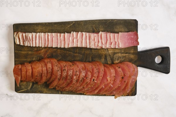 A wooden cutting board displays two types of cured meat. One side has thin slices of pink meat while the other has thicker slices of seasoned meat. This setup is ideal for meal preparation