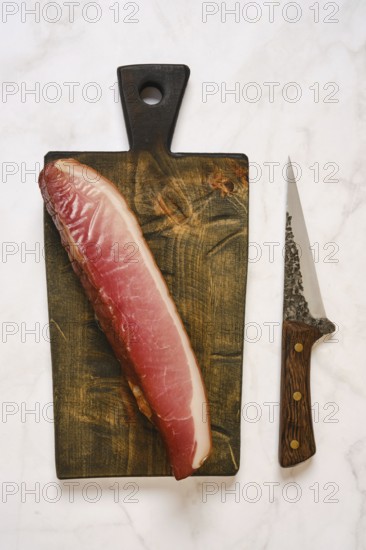 Cured parma ham on a wooden cutting board. A sharp knife is placed next to it. This scene is set in a kitchen, ideal for meal preparation or cooking activities