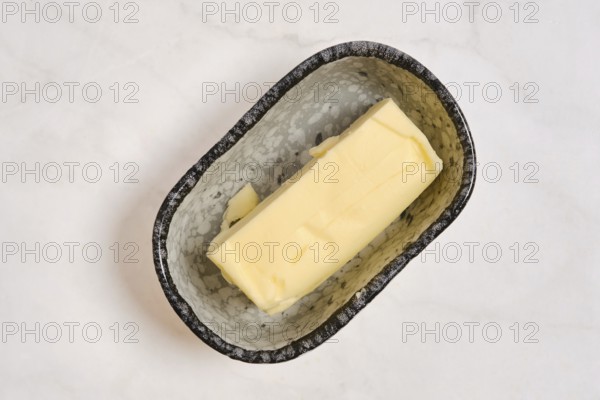 A piece of butter in a grey dish on a white countertop. This setting shows a kitchen environment with a focus on food preparation. The butter is smooth and unwrapped