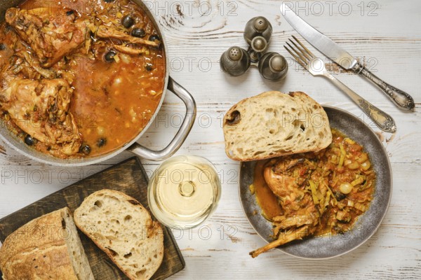 A rabbit stew with olives and vegetables sits in a pan next to a bowl. A piece of bread lies beside the bowl. A glass of white wine stands nearby. The setting shows a simple dining scene