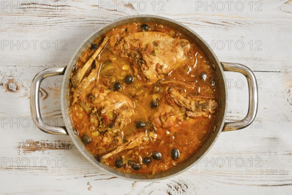 Rabbit legs are cooking in a pot filled with a sauce and olives. The pot sits on a wooden table. The scene shows preparation for a meal in a home kitchen during the day