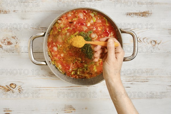 A hand sprinkles spice in a pot with chopped vegetables and a rich tomato base. Spices are added to enhance the flavor. The kitchen has a light wooden background