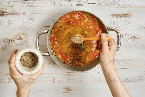 A person adds pepper to a pot of vegetables and tomatoes. The hands hold a wooden spoon and a mortar. Fresh ingredients are mixed as the meal is being prepared in a cozy kitchen