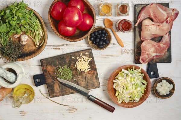 Fresh tomatoes, herbs, and meat are spread on a wooden table. There are bowls of spices and chopped vegetables nearby. A knife rests on a cutting board, ready for cooking