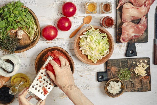 A person is grating tomatoes and chopping vegetables. Fresh herbs, spices, and rabbit meat are laid out on a wooden surface. The setting is a bright kitchen