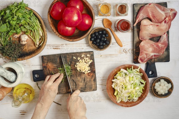 Hands are cutting rosemary on a wooden board. Fresh tomatoes, herbs, spices, and rabbit legs are arranged on the table. Various bowls hold chopped vegetables and ingredients ready for cooking