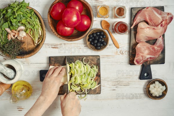 Fresh vegetables, meats, and spices are arranged on a table. Someone is chopping onions with a knife while tomatoes and other ingredients are nearby. This scene shows meal preparation in a kitchen