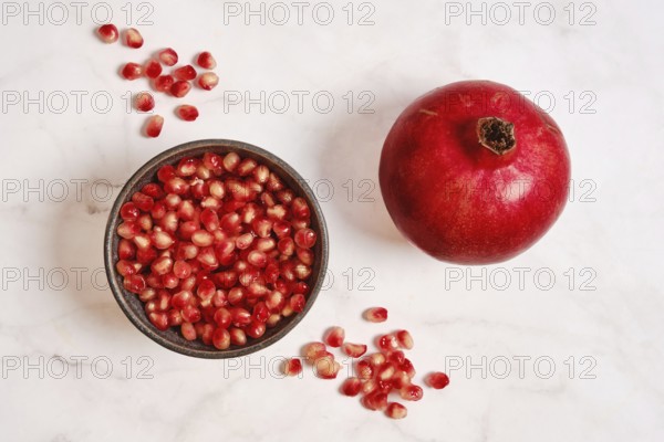 A round red pomegranate sits next to a bowl filled with bright red seeds on a white marble surface. The natural shine of the pomegranate stands out against the simple background