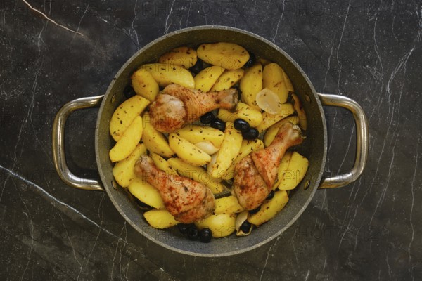Chicken drumsticks are cooking with potato wedges and olives in a pan on a dark kitchen counter. The food is seasoned and ready for serving during a family dinner time