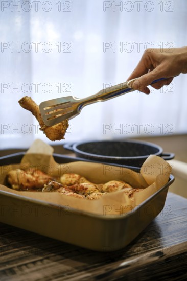 A hand lifts a piece of grilled chicken drumstick from a tray. The scene shows a cozy kitchen setup with natural light coming through the window