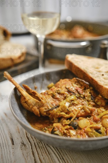 A plate features rabbit meat cooked with vegetables in sauce. Slices of bread rest next to the dish. A glass of wine stands nearby. The setting shows a warm kitchen atmosphere