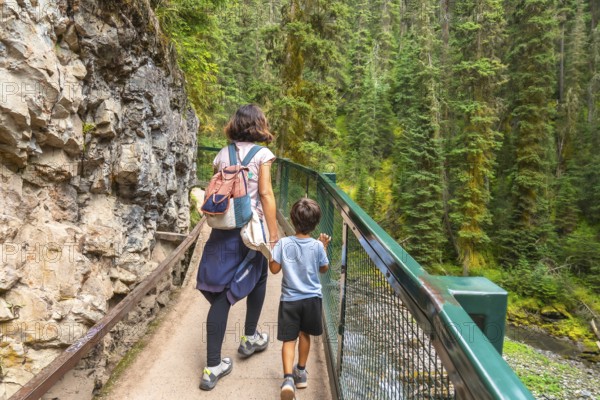 Mother and son holding hands are enjoying a hike along the scenic johnston canyon trail in banff national park, surrounded by lush green forest and towering rock formations