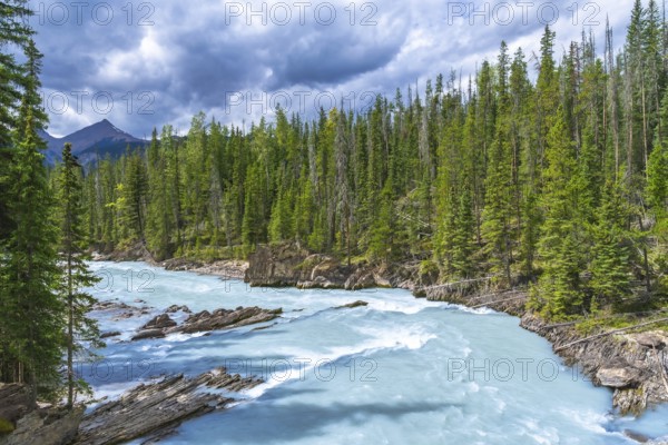 Turquoise water flows through the bow river natural bridge in banff national park, alberta, canada, surrounded by lush green pine trees and a cloudy sky