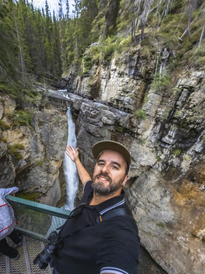 Photographer taking a selfie on a metal walkway, capturing the majestic johnston canyon waterfall cascading through the rugged cliffs of banff national park, alberta