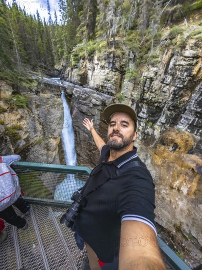 Tourist taking a selfie with outstretched arm at johnston canyon lower falls, showcasing the stunning waterfall, canyon, and lush forest in banff national park, alberta, canada, during a sunny day