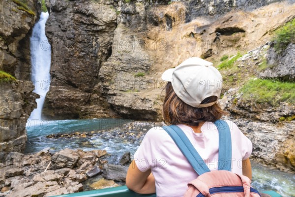 Female tourist wearing a backpack and baseball cap admiring a stunning waterfall cascading into turquoise water in johnston canyon, banff national park, alberta