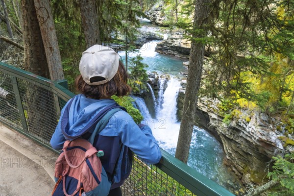 Female hiker wearing backpack and cap admiring cascading waterfalls and turquoise water of johnston canyon in banff national park, canadian rockies, alberta, canada, during summer vacation