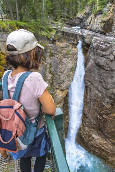Female tourist carrying a backpack and baby carrier, enjoying the stunning view of a waterfall cascading into the turquoise waters of johnston canyon in banff national park