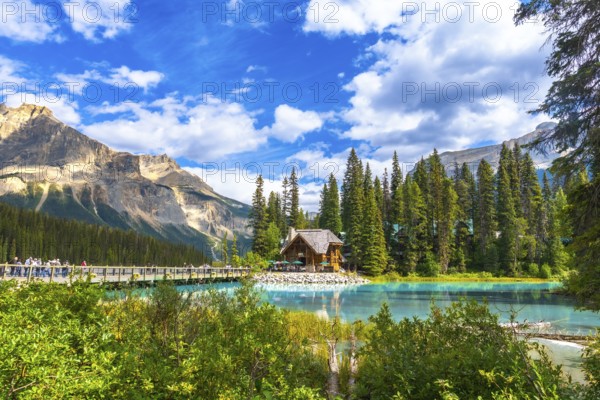 Wooden lodge reflecting on turquoise water with tourists walking on a bridge, surrounded by mountains and forest on a sunny day at emerald lake in yoho national park, canada