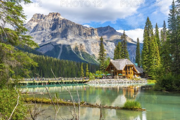 Wooden lodge reflecting in turquoise water on sunny summer day with tourists walking on bridge and mount burgess towering in background at emerald lake in yoho national park. British columbia. Canada
