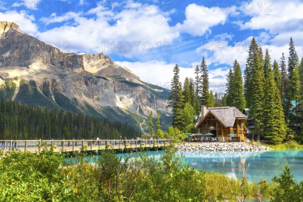 Wooden lodge sitting on the shore of a turquoise lake reflecting the surrounding lush vegetation and mountain range under a blue sky with clouds