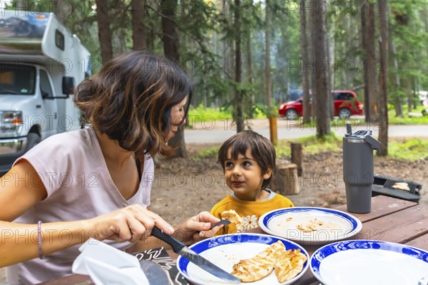 Mother giving grilled food to her child while having lunch at a campsite in the canadian rockies, banff national park, alberta, with their campervan parked in the background