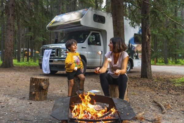 Mother and son roasting marshmallows over a campfire, enjoying quality time together during a camping trip in banff national park, with their campervan parked nearby in the serene forest