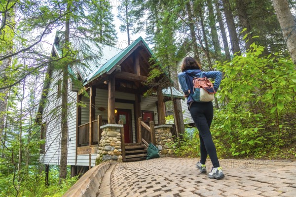 Female tourist with backpack walking up cobblestone path to a wooden cabin surrounded by lush green forest in banff national park, alberta, canada, enjoying a peaceful vacation getaway in nature