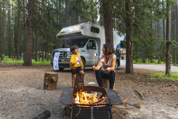 Mother and son roasting marshmallows over campfire near their camper van, enjoying a camping trip in the beautiful wilderness of banff national park, canadian rockies, alberta, canada