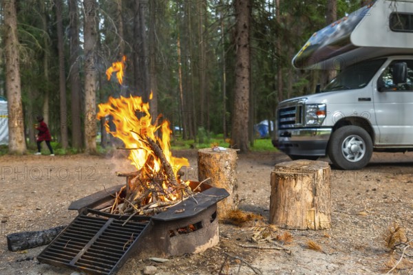 Warm fire glowing in a campsite fire pit beside a camper van in banff national park, alberta, surrounded by a coniferous forest and a tent, creating a cozy atmosphere